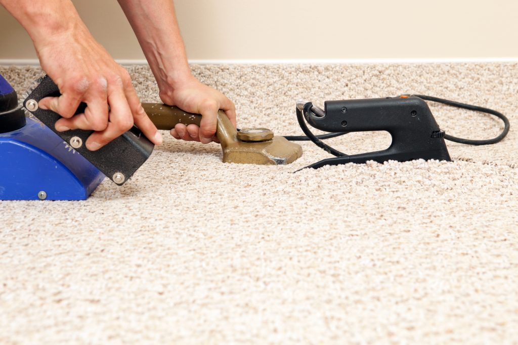 "A carpet installer is using a seam iron to join two sections of berber carpet in a bedroom at a house construction site. The left hand is holding a knee kicker carpet stretcher, and the right is operating a seam roller to hide the joint. The blue tool is a seam vacuum which extracts heat and pulls the seam tape up to the carpet. This image represents every component of the carpet seaming process."