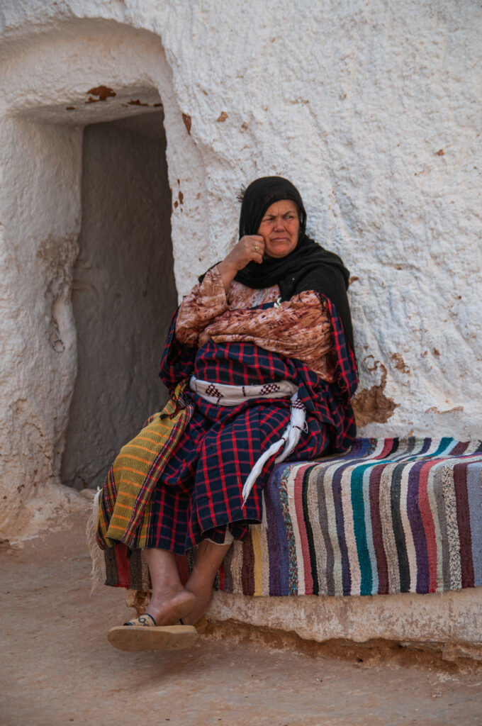 Old berber woman sitting on a bench covered with colorful rug in front of her traditional white house in Tunisia